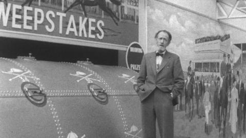 Fyfe Robertson, wearing a suit and bow tie, stands in front of the giants sweepstakes drum.