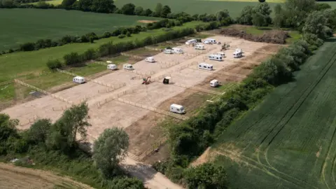 A drone bird's eye view of a field. It appears brown, as if hard standing has been laid over it. There are white caravans parked, dotted around the field, and at least one digger.