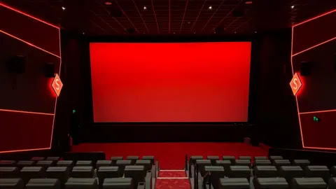 A view from inside a cinema room. There are rows of black leather seats and a red screen at the front. There is red branding down the sides