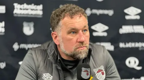 BBC/Matt Dean The photo shows a man looking past the camera. He's wearing a grey jacket with the Grimsby Town logo on. His microphone also has the Grimsby Town logo on it. He has grey hair and a grey beard, and is sitting behind a board with different logos on.