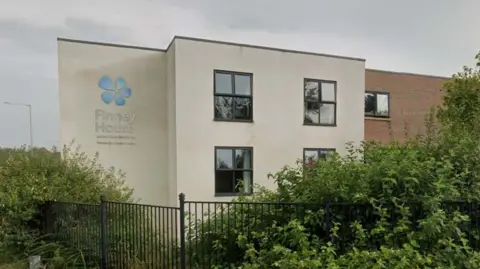 Street view image of Finney House, a large cream rendered building with black-framed windows and a blue flower logo. there are railings and bushes in front