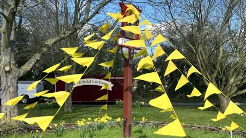 Helen Mulroy/BBC Thriplow's village sign is in the middle and on a a section on grass. There are several trees behind the sign, and on the sign there is yellow triangular bunting.