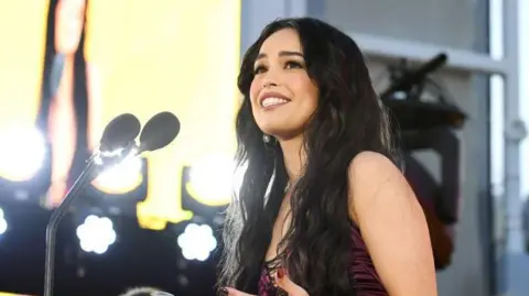 Getty Images Valkyrae at a gala event in LA in 2024. She has long dark hair worn loose and wears a burgundy dress. She smiles and looks up as she delivers a speech on stage. 