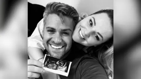 Russo family A black and white image of Daniel and Bryony Russo holding a printed baby scan showing Emmy in Bryony's womb. They are both hugging and smiling.
