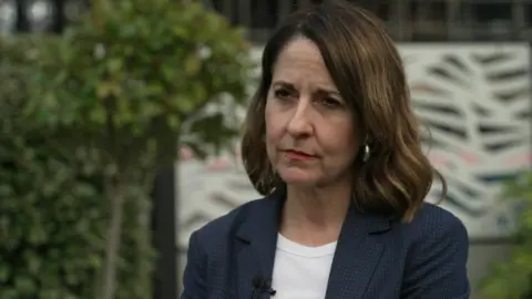 BBC Close-up of Pensions Secretary Liz Kendall in a blue jacket and white top with trees and bushes in the background