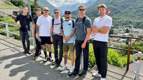 Phillip Norton/BBC A group of seven men are standing in front of a metal fence which looks out on lots of buildings, trees and mountains below. Some are wearing backbacks and holding water bottles with serious expressions on their faces. The sun is out and it looks like a hot day.