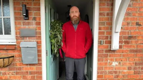 Matty Williams standing in his doorway, he is bald and has a ginger beard. He is wearing a red fleece. The front door is turquoise and nicely contrasts to the red brick of the exterior wall of the house. There is a Christmas wreath on the door.