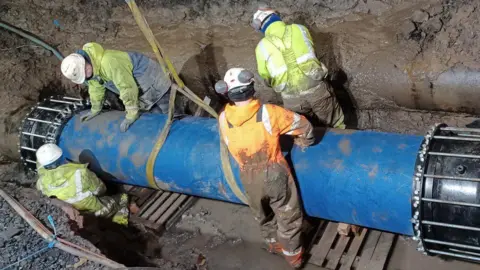 Scottish Water Four engineers wearing muddy fluorescent waterproofs and white helmets as they work to repair the burst water main on Pollokshaws Road in Glasgow. They are in a ditch securing a large blue pipe section, which has the yellow sling used to lower it still attached.