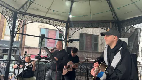 Four men play instruments under a bandstand. On the right is Marty, and he wears a black and grey changing robe and a baseball hat. He plays the guitar. There is a bald man singing and a bearded man on a guitar.