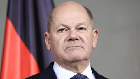 Reuters A headshot of Germany's Chancellor Olaf Scholz as he appears expressionless photographed in front of a red and black flag