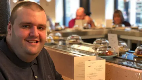 Cancer Research UK A man with a buzz cut hair style wearing a dark grey polo top is pictured sitting in a sushi restaurant.