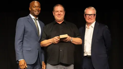 West Midlands Combined Authority Chris Bruce with actor Nicholas Bailey and West Midlands mayor Richard Parker. Chris has short dark hair and wears a black shirt and grey trousers while holding a box. To his right is Richard Parker in a dark blue suit and white shirt, he wears black glasses. Nicholas Bailey is on the left and has a shaved head and wears a lighter blue suit, a white shirt and olive-green tie.