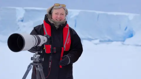Sue Flood A woman standing behind a large lensed camera atop a tripod. She is wearing a thick black coat, black gloves, a neutral-coloured scarf and glasses with yellow lenses and rims.
She had flowing silver hair and is smiling. In the background is a large sheet of snow and a white background with the ground entirely covered in snow.
She also has a red water storage device around her neck with a black and red strap