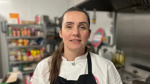 A woman with long dark hair in a white top and black apron facing the camera with shelves of catering supplies behind her