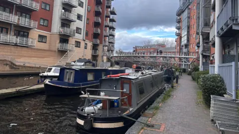 It is a straight canal with several long narrowboats and small vessels moored along both sides, next to paved walkways. Modern multi‑storey apartment buildings line both banks, built from red brick and light‑coloured materials, many with balconies facing the water. In the background, a pedestrian bridge crosses the canal beneath a sky with scattered clouds.