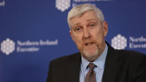 O'Dowd has grey hair and a grey beard. He is wearing a black suit and light blue shirt with a red chequered tie.