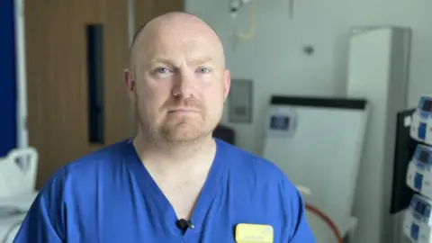 Dr Scott Grier Consultant in Intensive Care Medicine. He has very close cropped hair and stubble. He has blue eyes and is wearing blue scrubs.
