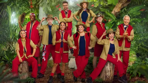 ITV 10 people can be seen wearing various outfits of red and khaki green, with some wearing large khaki hats. They are posed in front of a jungle backdrop as part of the announcement for the new cast of I'm a Celebrity...Get Me Out of Here!