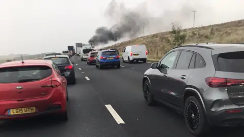 A queue of stationary traffic. Black smoke can be seen rising in the distance