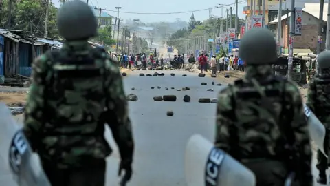 AFP Two Kenyan riot police officers with their backs to the camera holding shields and people standing near a roadblock during a protest in Kisumu after the election results were announced on August 9, 2017