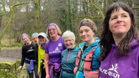 A group of women in colourful running clothes line up along a bridge in the countryside.