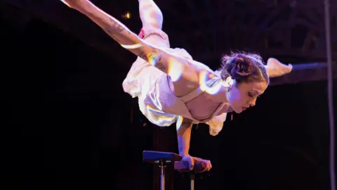 Andre Pattenden A performer on a stage balancing with one hand on a beam. There are dreamy refracted lights shining on her and she is wearing a white costume with her hair in bunches.