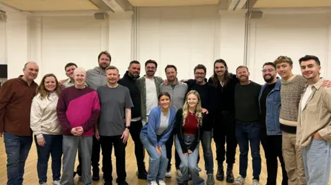 Luke Wilby 17 people stand in a line in a rehearsal room. They are wearing casual clothes and smiling at the camera