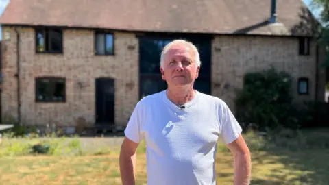 Rob Taylor wears a white T-shirt and has his hand on his hips. He stands in his back garden and behind him is the barn conversion he rents from HS2. 