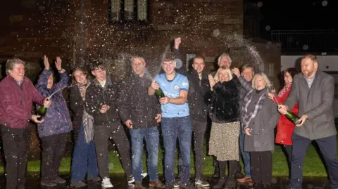 National Lottery James Clarkson surrounded by family and friends, spraying champagne in a garden.