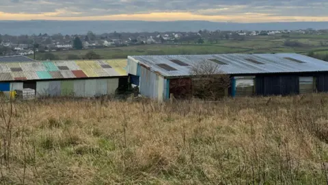Corrugated iron farm buildings on empty green land overlooking hills with houses in the distance