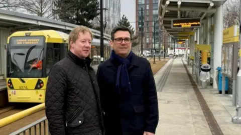 Cambridgeshire and Peterborough Combined Authority Two men, wearing thick winter coats stand on a tram platform. A yellow tram is visible in the background. 
