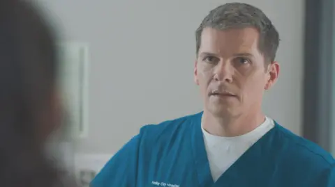 Nigel Harman playing Dr Max Christie in Casualty. He wears blue-green scrubs over a white T-shirt. He has short, slightly greying hair and looks seriously at another character who is blurred on the left foreground.
