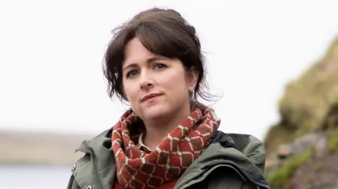 A young woman with dark hair stands on a rocky outcrop, next to open water. She is wearing a green jacket and a red scarf. 