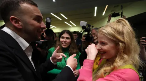 EPA Green Party leader Zack Polanski speaks to Green Party MP Hannah Spencer who smiles as photographers take pictures around them. A party worker stands in the background.