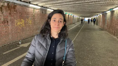 A woman in a black jacket looking at the camera inside an underpass. People walk and cycle behind her and a small pigeon stands behind her on the left.