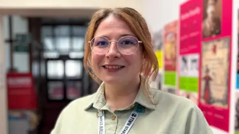 Isabel is wearing a pale green shirt and FTWW lanyard. She is stood in a corridor with brightly coloured posters on the wall behind her. She has round glasses and her brunette hair is tied back with some strands framing her face.