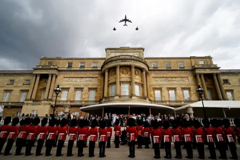 Reuters Veterans and members of the armed forces gather outside Buckingham Palace and gaze up to watch RAF aircraft fly over the palace against a cloudy grey sky.
