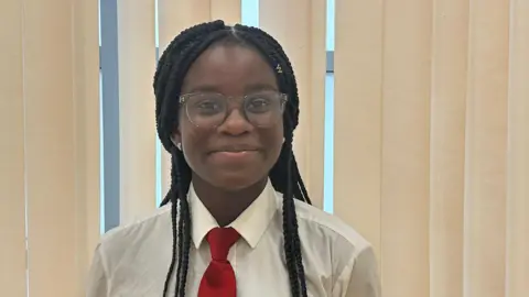 Malcolm Ring/BBC A girl with black braided hair and glasses. She is wearing her school uniform, which is a white shirt with a red tie. She is stood in front of some cream-coloured window blinds.