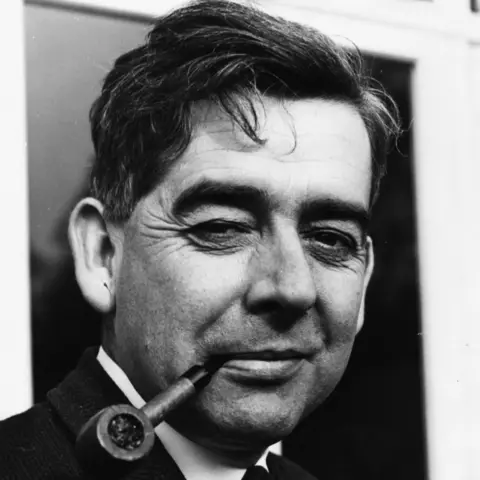 Getty Images Black-and-white close-up portrait of a middle-aged man wearing a suit and tie, looking slightly to camera, holding a pipe in his mouth, with a building façade blurred in the background.