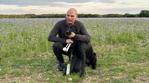 BBC correspondent Sean Dilley pictured in a field on a bright day, crouched down with his arms around his black guide dog Shawn