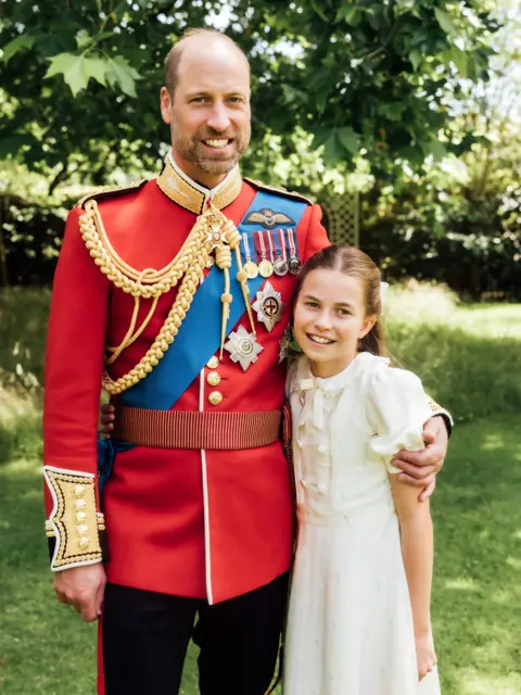 MATT PORTEOUS / KENSINGTON PALACE Prince William is wearing the scarlet dress uniform of the Welsh Guards, of which he is colonel, and has his arm around Charlotte, who is wearing a cream dress. 