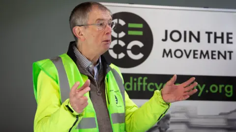 Eva Marloes Chris Roberts stands at an event with a viz vest and jacket open over his grey jumper. he is holding his hands up in the air at a he speaks in front of a campaign sign on the wall behind him that say join the movement. 