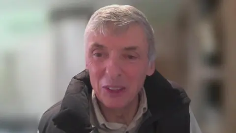 Jeremy Barrell is pictured in a video call. He has light grey hair and wears a padded gilet over a light, collared shirt.