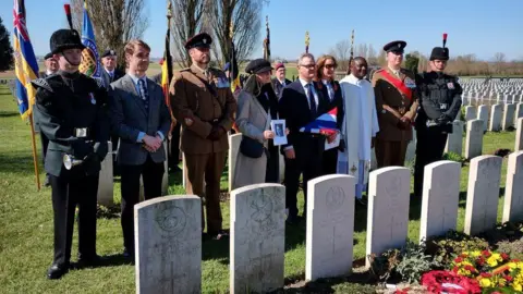 Eight people, including civilians in smart clothes, a priest in white robes s and men in military uniforms stand in a line in front of a row of white headstones. Behind them are military personnel holding flags. Rows of gravestones can be seen beyond. The sky is blue.