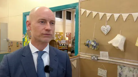 Stephen Kinnock in a blue suit and tie stands indoors, slightly turned to one side, with a small microphone clipped to the jacket. The setting appears to be a community or classroom space, with light‑coloured walls and a doorway behind them leading to another room containing shelves, children’s items, and a high‑visibility jacket hanging on the wall.