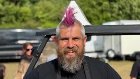 Spudman smiles at the camera wearing a black T-shirt and black apron. He has  a pink mohawk and a long beard. 