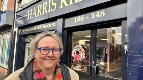 Kim Harris. She has blonde hair, wearing glasses, a beige coat with black collar. A blue shop-front for Harris Kilts is behind her.