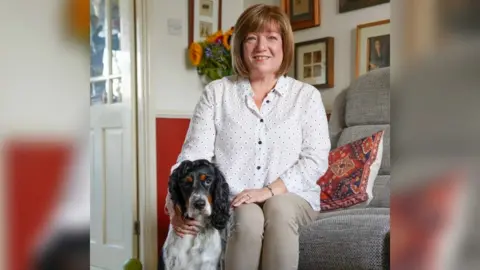 Hearing Dogs Watson is sitting by Jeannette Godsell's legs. Jeannette is sitting on the sofa and smiling at the camera. She is wearing a white blouse and beige jeans.