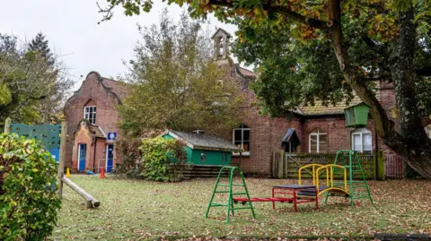 View of Corpusty Primary School. It is an old brick building with brightly coloured childrens play equipment on the grass in front of it.