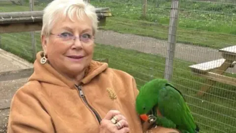 A woman with short blonde hair and glasses. She is wearing a brown coat and has rings on her finger. There is a green parrot perched on her left arm and there is a fenced enclosure in the background.
